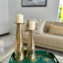 Load image into Gallery viewer, Two gold candle holders with white candles on a green tray in a living room setting.
